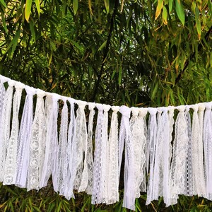 May include: A white lace garland with multiple strands of varying lace patterns. The garland is suspended against a backdrop of green foliage. The lace appears to be made of cotton or a similar fabric, and is suitable for weddings or other celebrations.