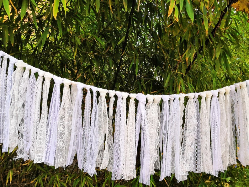 May include: A white lace garland with multiple strands of varying lace patterns. The garland is suspended against a backdrop of green foliage. The lace appears to be made of cotton or a similar fabric, and is suitable for weddings or other celebrations.