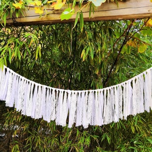 May include: A white lace banner with a scalloped edge hangs against a backdrop of green bamboo. The banner is made of strips of white lace and fabric, creating a delicate and textured appearance. The banner is suspended from a white rope.