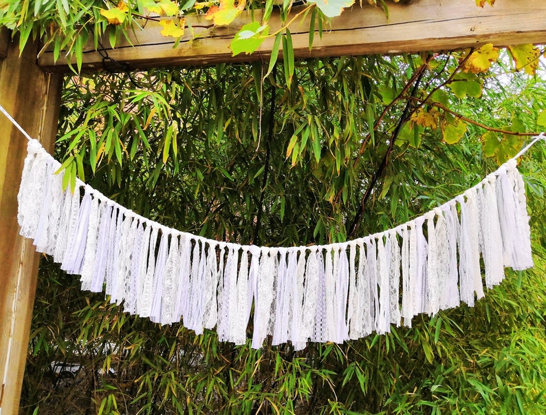 May include: A white lace banner with a scalloped edge hangs against a backdrop of green bamboo. The banner is made of strips of white lace and fabric, creating a delicate and textured appearance. The banner is suspended from a white rope.