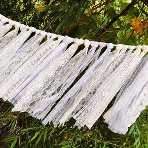 May include: A white lace garland with various lace patterns and textures. The garland is strung with knotted white fabric and hangs against a backdrop of green foliage. Suitable for weddings or other celebrations.