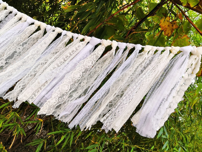 May include: A white lace garland with various lace patterns and textures. The garland is strung with knotted white fabric and hangs against a backdrop of green foliage. Suitable for weddings or other celebrations.