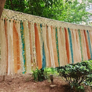May include: A decorative hanging garland featuring a wide, cream-colored crocheted lace header with numerous vertical fabric strips attached. The strips consist of various textures and colors, including solid cream, orange, and teal blue lace.