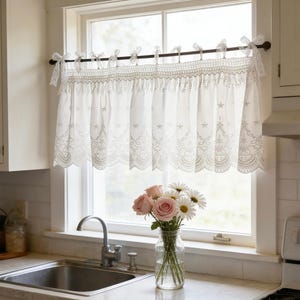 May include: White lace kitchen curtains with a scalloped edge and star embroidery hang above a kitchen sink. A bouquet of pink roses and white daisies sits in a glass vase on the countertop.