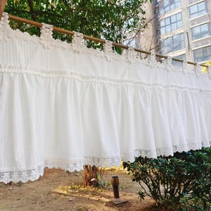 May include: White lace-trimmed curtain hanging outdoors. The curtain is made of a soft, flowing fabric and is suspended from a wooden rod. The top edge features decorative floral accents, and the bottom edge has a delicate lace trim. The curtain is a bright white colour.