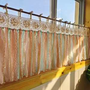 Może przedstawiać: Garnitur okienny wykonany z koronki szydełkowej z rzędem pasków tkaniny zwisających poniżej. Paski tkaniny są w odcieniach brzoskwiniowego, różowego i miętowego.