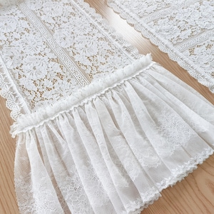 Pode incluir: Caminho de mesa de renda branca com padrões florais e borda com babados. O caminho de mesa é feito de tecido delicado e transparente, projetado para uso decorativo. O design de renda intrincado adiciona elegância.