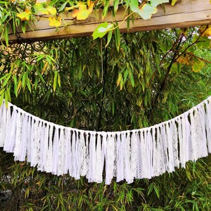 May include: A white lace and fabric banner hangs against a backdrop of green foliage. The banner is made of various white lace and fabric strips, creating a decorative, textured effect. The banner is suspended from a white string.