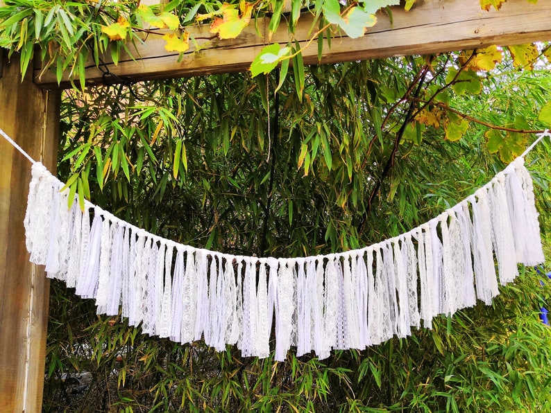 May include: A white lace and fabric banner hangs against a backdrop of green foliage. The banner is made of various white lace and fabric strips, creating a decorative, textured effect. The banner is suspended from a white string.