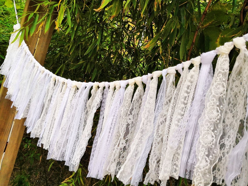 May include: A white lace garland with different lace patterns and textures. The garland is strung with white ribbon and has delicate floral accents. The background includes green foliage and a wooden post.