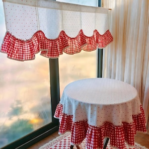 May include: A white and red gingham-patterned valance and matching tablecloth with a ruffled edge. The valance has a white dotted fabric with a red gingham ruffle. A round table is covered with a matching tablecloth.