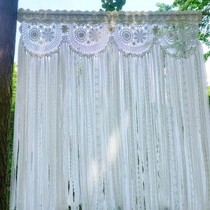 May include: White crochet curtain with a scalloped top and long fringe. The curtain is hanging from a wooden dowel rod.