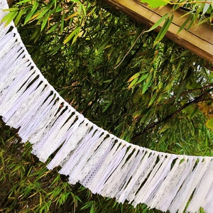 May include: A white lace garland with a ruffled design hangs against a backdrop of green foliage and a wooden structure. The garland is made of layered fabric and lace, creating a textured, decorative element.