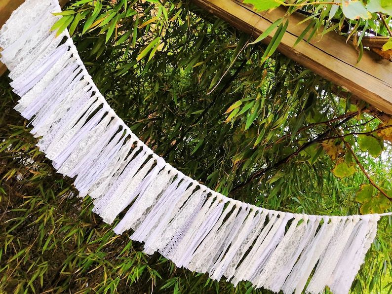 May include: A white lace garland with a ruffled design hangs against a backdrop of green foliage and a wooden structure. The garland is made of layered fabric and lace, creating a textured, decorative element.