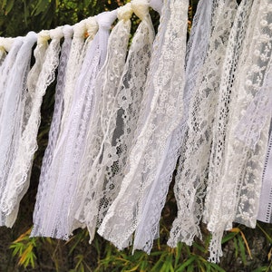 May include: A white lace garland featuring a variety of lace designs. The garland is constructed from different types of white lace, producing a textured and layered appearance. The lace is draped and suspended, set against a natural backdrop.