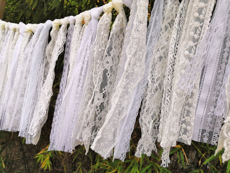 May include: A white lace garland featuring a variety of lace designs. The garland is constructed from different types of white lace, producing a textured and layered appearance. The lace is draped and suspended, set against a natural backdrop.