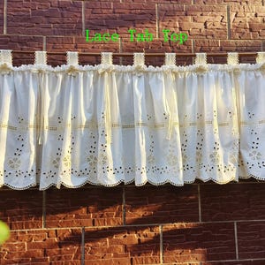 May include: Cream-colored lace tab top curtain with floral and dot cutouts. The curtain is hanging on a brown rod against a brick wall. The text "Lace Tab Top" is visible in green.