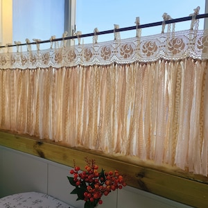 May include: A white lace curtain with a fringe of thin fabric strands hangs from a window. The curtain is tied back on each side with a white ribbon. The curtain is in front of a window with a view of a blue sky.