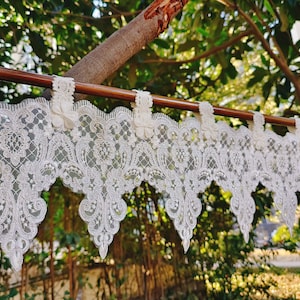 Pode incluir: Valance de renda branca com borda festoneada e delicado padrão floral. A valance está pendurada em uma haste de madeira.