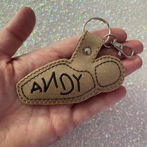 May include: A gold glittery keychain shaped like a boot with the name ANDY embroidered in black thread on the side. The keychain features a silver metal split ring and a lobster claw clasp attached with a silver rivet at the top of the boot design.