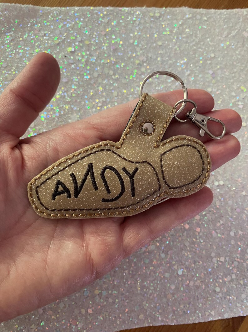 May include: A gold glittery keychain shaped like a boot with the name ANDY embroidered in black thread on the side. The keychain features a silver metal split ring and a lobster claw clasp attached with a silver rivet at the top of the boot design.