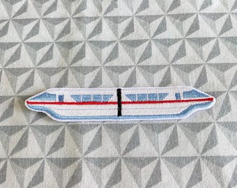 May include: A white, blue, and red embroidered patch of a monorail train. The patch is on a white background with a gray geometric pattern.