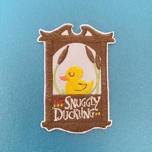 May include: Embroidered patch featuring a yellow duckling inside a wooden frame with the text "The Snuggly Duckling".