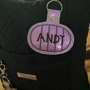 May include: A glittery purple oval-shaped keychain tag featuring the name ANDY embroidered in black thread. The tag has vertical purple stripes, a green stitched border, and a silver metal snap button, attached to a silver split ring and lobster clasp.