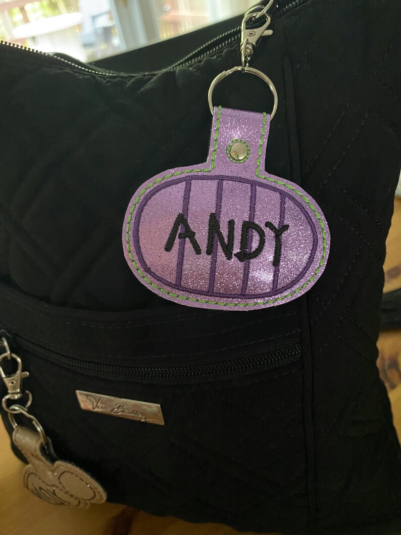 May include: A glittery purple oval-shaped keychain tag featuring the name ANDY embroidered in black thread. The tag has vertical purple stripes, a green stitched border, and a silver metal snap button, attached to a silver split ring and lobster clasp.