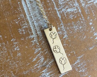May include: Gold-colored pendant necklace with three etched Mickey Mouse balloon designs on a rectangular bar. The necklace has a delicate chain and is displayed on a textured, light brown surface.