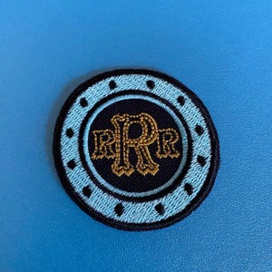 May include: A circular embroidered patch with a light blue border and a navy blue center. The center features the gold-colored letters "RRR" in an ornate font. The patch has a black outer border and is set against a blue background.