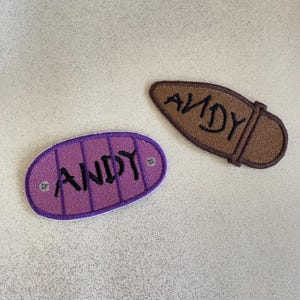 May include: Two embroidered patches with the name "ANDY". One is purple with a stitched border and vertical lines, the other is brown with a strap design. Both have black lettering.