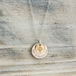 Puede incluir: Un collar de cadena de plata con un colgante de plata y oro. El colgante tiene las palabras "Off to Neverland" grabadas y una estrella de brújula dorada en el centro.