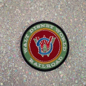 May include: A circular embroidered patch featuring a red center with a blue and green WD logo. The border is green with the white text WALT DISNEY WORLD RAILROAD. The patch has a black outer edge and is stitched with detailed thread work.