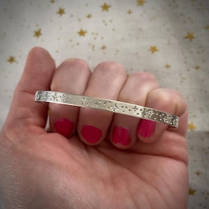 May include: A silver bangle bracelet with a repeating pattern of small stars and dots.