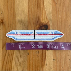 May include: A white, blue, and red embroidered patch of a monorail train. The patch is approximately 2 inches long and is measured against a ruler.
