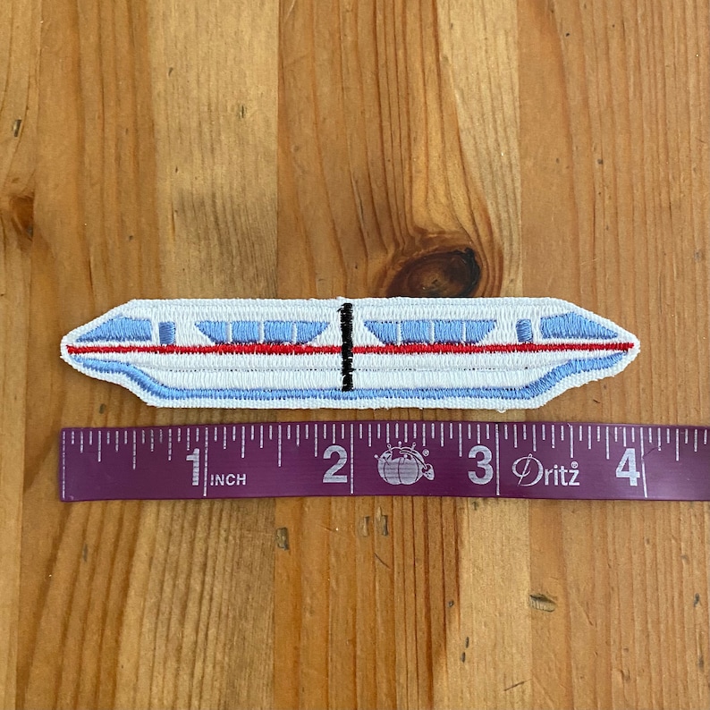 May include: A white, blue, and red embroidered patch of a monorail train. The patch is approximately 2 inches long and is measured against a ruler.