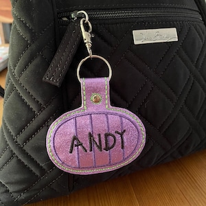 May include: A purple glittery oval keychain with the name ANDY embroidered in black thread. The keychain features vertical stitching, a lime green border, and a silver metal snap hook attached to a black quilted backpack with a silver logo plate.