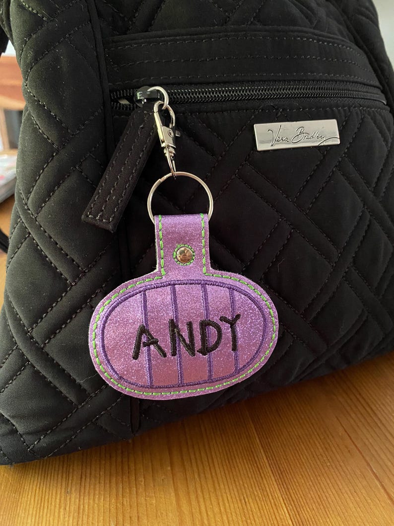 May include: A purple glittery oval keychain with the name ANDY embroidered in black thread. The keychain features vertical stitching, a lime green border, and a silver metal snap hook attached to a black quilted backpack with a silver logo plate.