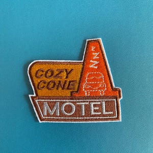 May include: A brown and orange embroidered patch with the text "Cozy Cone Motel" in white. The patch features a stylized orange traffic cone with a white stripe and a car silhouette.