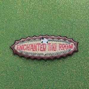 May include: Embroidered patch with the text "ENCHANTED THE ROOM" in red, surrounded by a brown and beige design. A small white flower is at the top. The patch is on a green background.