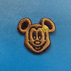 May include: Embroidered patch featuring a smiling cartoon mouse face with large ears. The patch is primarily brown with dark brown and yellow accents, set against a blue background. The patch is likely for clothing or accessories.