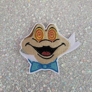 May include: An embroidered patch featuring a tan frog character with a wide, black-outlined smile and a blue bowtie. The frog has prominent eyes with red and yellow swirling hypnotic patterns. The patch has a white border and a white fabric backing.