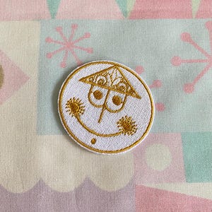 May include: A white embroidered patch with a gold outline. The patch features a stylized smiley face with a house-shaped hat and two eyes. The house has a decorative design on the roof.