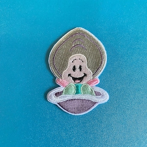 May include: Embroidered iron-on patch featuring a cartoon character with a smiling face, wearing a green and purple outfit, sitting in a flying saucer. The character has a light brown shell and pink cheeks.