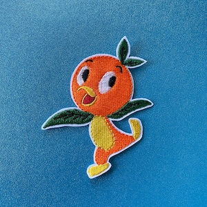 May include: Orange bird embroidered patch with a yellow beak, white eyes, and green leaves. The bird is standing on one leg and has a yellow body.