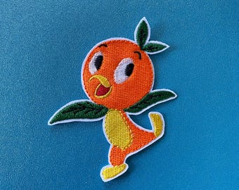 May include: Orange bird embroidered patch with a yellow beak, white eyes, and green leaves. The bird is standing on one leg and has a yellow body.