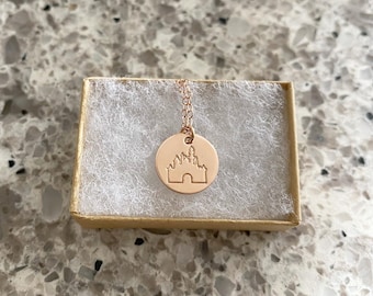 May include: A rose gold necklace with a round pendant featuring a silhouette of Cinderella's castle. The necklace is in a small, brown gift box.