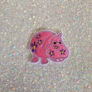 May include: Pink embroidered iron-on patch of a cartoon hippopotamus with purple flowers and yellow centers.