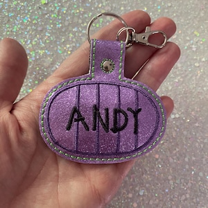 May include: A purple glittery oval keychain featuring vertical dark purple stripes and the name ANDY embroidered in black thread. The keychain is attached to a silver metal split ring and a lobster clasp, with bright green stitching along the edges.